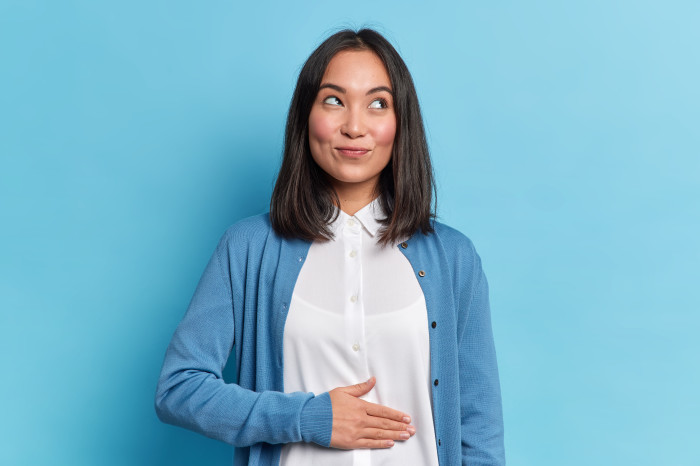Pleased cheerful Asian woman keeps hand on belly feels full after delicious dinner dressed casually stands thoughtful against blue background. Happy young female finds out about her pregnancy